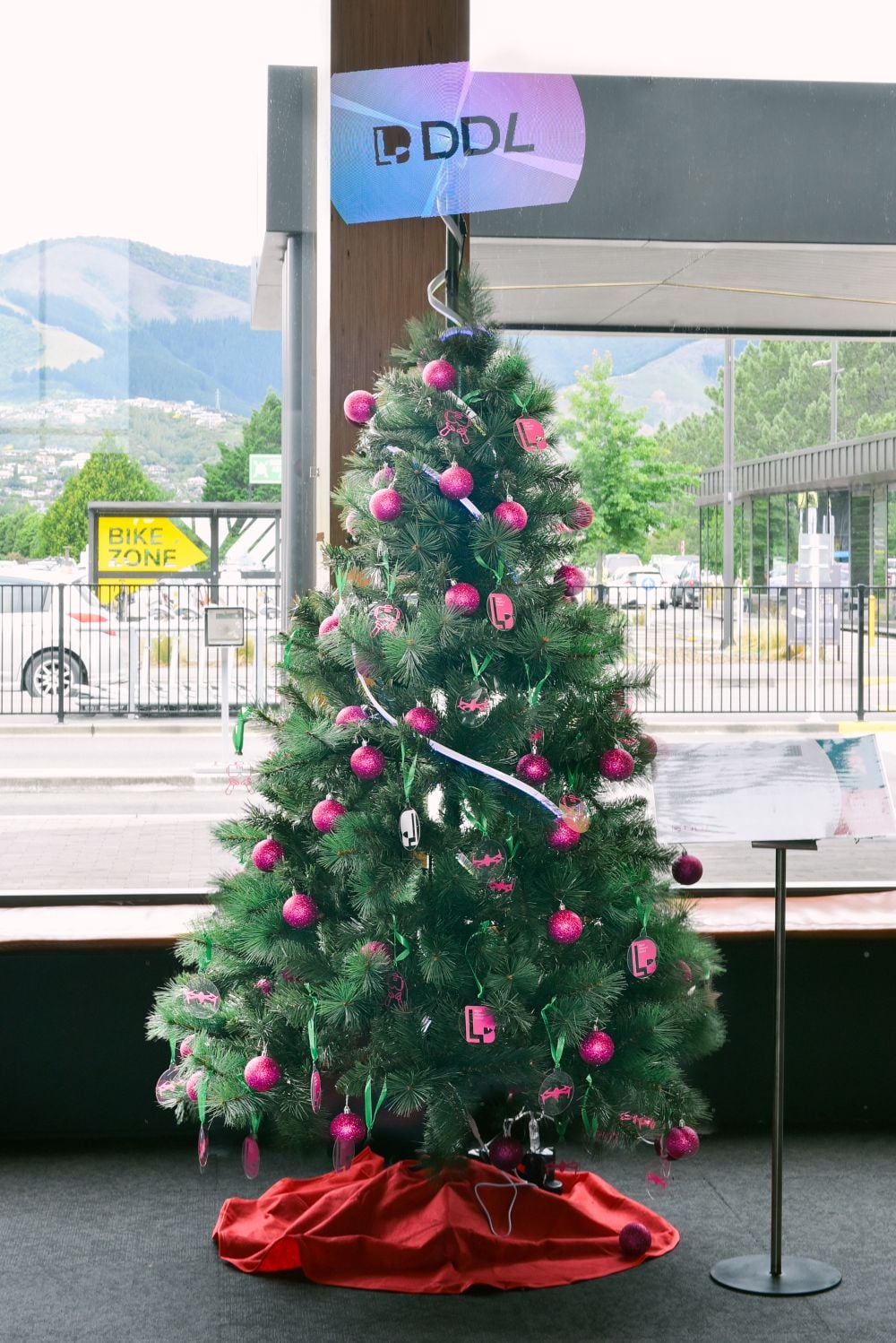 Christmas tree in the airport terminal, with pink and silver decorations and a digital tree topper displaying the DDL logo. Sponsored by Digital Diligence Ltd for the 2025 Nelson Airport Trees of Remembrance campaign.